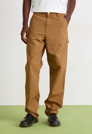 Person wearing loose brown pants with a white shirt tucked in, standing on a green floor, hands in pockets, and black shoes with yellow stitching.