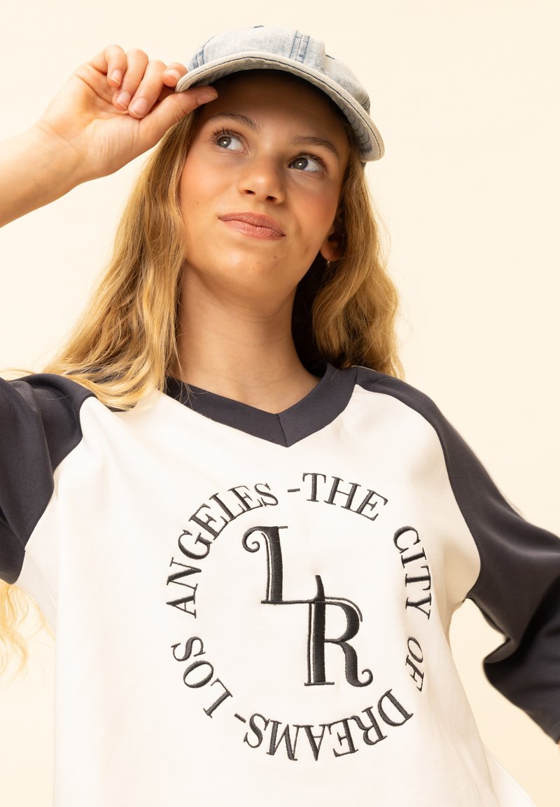 Witte en marineblauwe top met lange mouwen en een V-hals, met de tekst "Los Angeles - The City of Dreams" in een cirkelvormig ontwerp. Lichtblauwe denim pet.