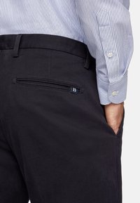 Dark gray corduroy trousers with a single back pocket featuring a hidden zipper and branded accent in navy blue. Striped shirt visible.