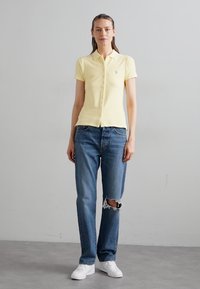 Yellow polo shirt with a collar, button-up front, paired with blue distressed jeans and white sneakers. Simple design, casual look.