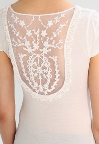 Biała bluzka z krótkim rękawem, z przezroczystym siatkowym tyłem z haftem kwiatowym i koronkowym wykończeniem, podkreślająca skomplikowane wzory i faktury.