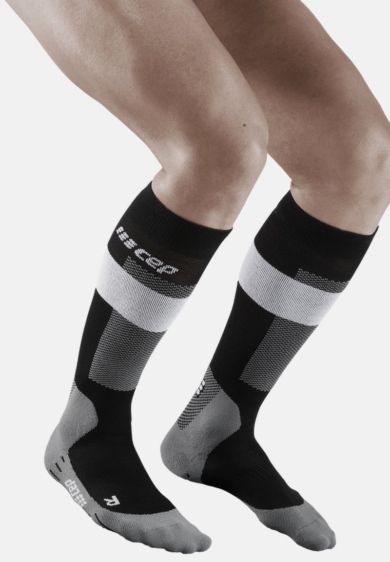 Calze a compressione in nero, grigio e bianco con motivi geometrici. Presentano un tallone e una punta rinforzati. Altezza fino al polpaccio con dettagli del logo.