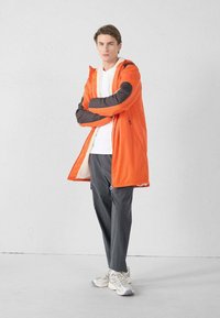 Jonge man staand met gekruiste armen, draagt een oranje jas met zwarte elleboogstukken, een wit overhemd, grijze broek en witte sneakers.