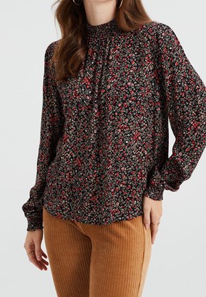 Blouse noire à motifs floraux avec des manches longues, encolure froncée, petites fleurs rouges et crème, associée à un pantalon marron côtelé.