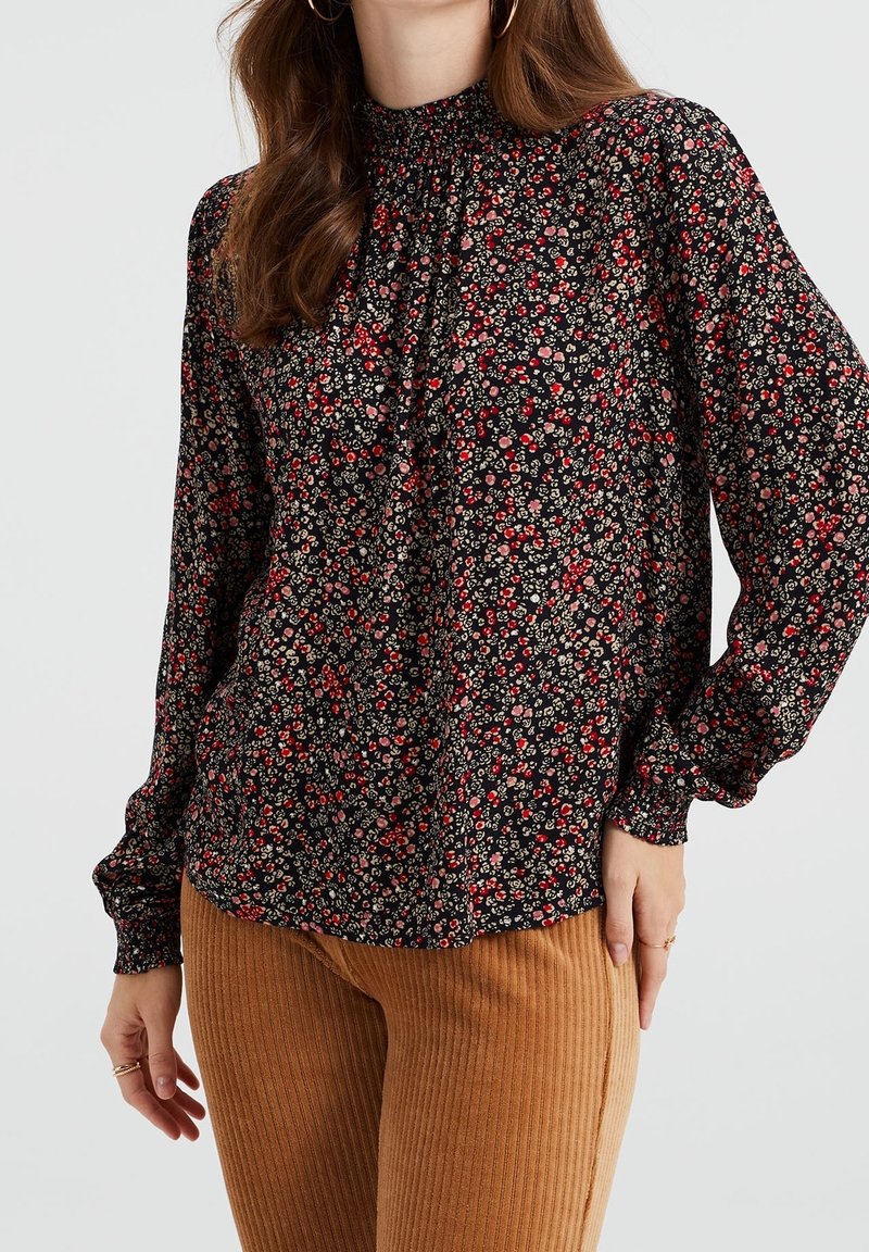 Svart blommig blus med långärmar, samlad halsringning, små röda och krämfärgade blommor, matchad med ribbade bruna byxor.