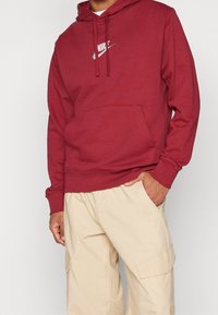 Röd huvtröja i tyg med en logotyp på bröstet, med en känguruficka och ribbade ärmslut, i kombination med beige cargobyxor.