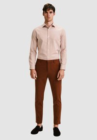 Camicia rosa chiaro a maniche lunghe con colletto bottonato, abbinata a pantaloni marroni aderenti e mocassini neri. Design semplice, texture liscia.