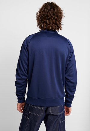 Homme aux cheveux bouclés portant une veste bleu marine unie à manches longues et un jean foncé, vu de dos sur un fond uni.