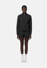 Felpa nera con zip, maniche lunghe e polsini a costine, abbinata a pantaloni corti neri. Il modello indossa sneakers alte bianche e calzini alla caviglia.