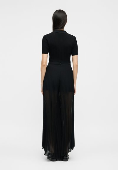 Γυναίκα με μακριά σκούρα μαλλιά, που φοράει μαύρο τοπ με κοντά μανίκια και διάφανο, πιετενέ παντελόνι φαρδιάς γραμμής, στέκεται γυρισμένη προς τα πίσω σε λευκό φόντο.