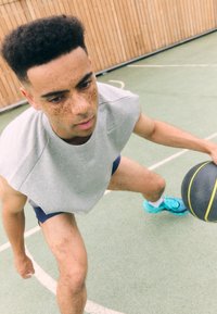 Grå ermeløs hettegenser, marineblå shorts, atletiske joggesko i aqua. Spilleren dribler en basketball på en teksturert grønn bane med hvite linjer.