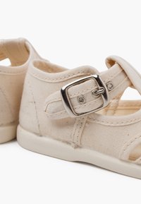 Beige canvas schoenen met een gespsluiting, open zijkanten en een witte rubberen zool, met stikdetails en een gladde textuur.