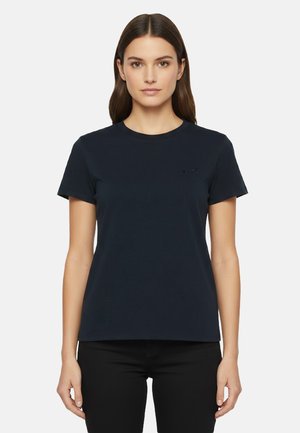 Donna con capelli castani lisci che indossa una maglietta a maniche corte blu scuro semplice e pantaloni neri, in piedi davanti a uno sfondo bianco uniforme.