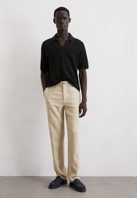 Polo noir à côtes, pantalon beige ajusté et mocassins foncés portés par un mannequin sur un fond neutre. Design simple et épuré.