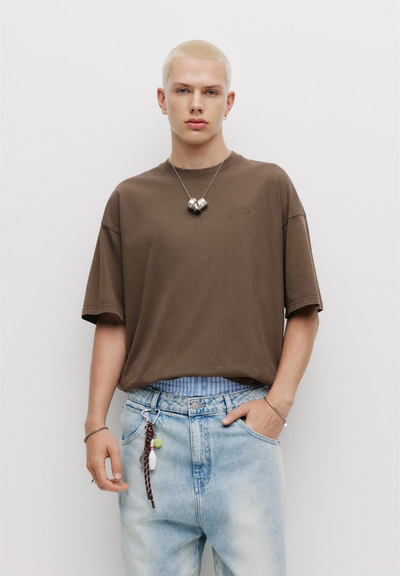 Hnědé oversized tričko s krátkým rukávem a kulatým výstřihem, spárované se světle modrými džínami s výdrží, které mají pruhovaný pas a klíčenku.