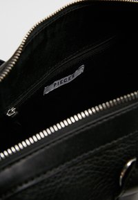 Framkant av en svart läderväska med ett fack med dragkedja och en vit etikett med texten "PIECES" inuti väskan.