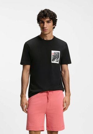 Junger Mann trägt ein schwarzes T-Shirt mit einem Palmenmotiv auf der Brusttasche und pinke Shorts mit Kordelzug, steht vor weißem Hintergrund.