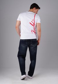Man wearing white T-shirt with red vertical text on back, dark jeans, and white sneakers, standing against gray background, facing away.