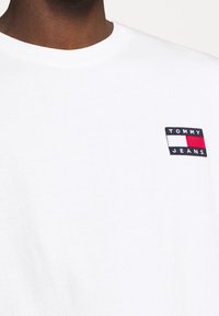 Bílá bavlněná tričko s malou obdélníkovou nášivkou s textem "TOMMY JEANS" a červenými/modrými akcenty na levé části hrudi.