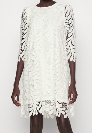 Witte knielange jurk met een ingewikkelde kanten overlay met bladmotief en driekwart mouwen, gedragen door een persoon met een donkere huidkleur die staat tegen een witte achtergrond.