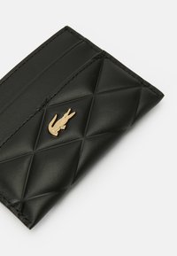 Portafoglio in pelle nera trapuntata con emblema di coccodrillo dorato e bordi cuciti su uno sfondo bianco.