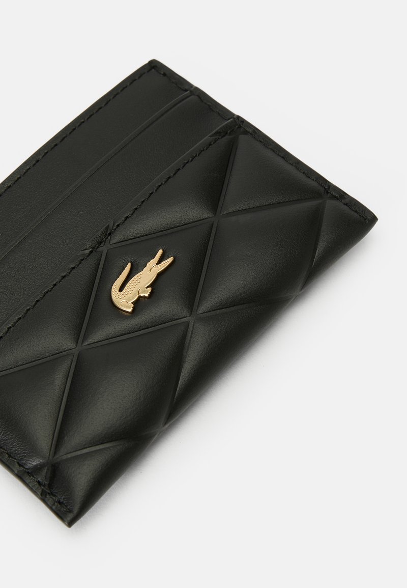 Porte-cartes en cuir matelassé noir avec emblème crocodile doré et bords surpiqués sur fond blanc.