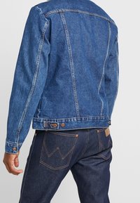 Džínová bunda v středně modré barvě s kontrastním prošitím, knoflíkovými detaily a klasickým střihem. Kombinována s tmavě modrými džínami s dekorativními kapsami.