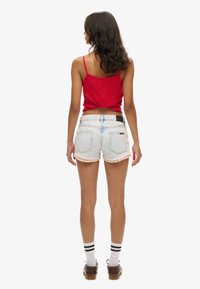 Top tipo tank rojo ajustado combinado con pantalones cortos de mezclilla de lavado claro, con un dobladillo deshilachado y un borde de encaje. La modelo lleva calcetines blancos con rayas negras y zapatos marrones.