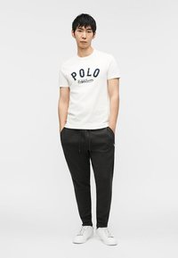 Jeune homme portant un t-shirt blanc Polo Ralph Lauren, un pantalon de jogging noir et des baskets blanches, debout les mains dans les poches devant un fond gris.