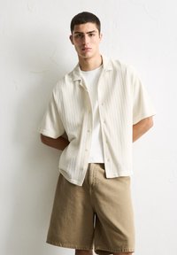 Cream button-up shirt with a textured pattern, paired with khaki knee-length shorts. Model stands against a plain textured background.
