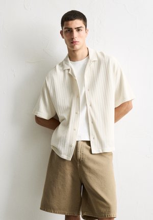 Chemise crème à boutons avec un motif texturé, associée à un short beige à hauteur des genoux. Le modèle se tient devant un fond texturé uni.