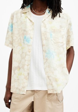 Uomo che indossa una camicia a maniche corte beige con motivi floreali sopra una maglietta bianca a coste, con pantaloni cargo beige e un accessorio a catena.