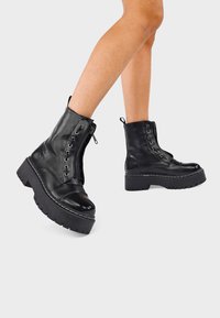 Botas de cuero negras hasta el tobillo con un acabado texturizado, con cordones en la parte frontal, cremallera lateral y suela de plataforma gruesa. Presenta acentos de costura blanca.