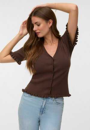 Kvinde med langt brunt hår iført en tætsiddende brun knapbluse med flæsekant og lyseblå jeans, poserer med armene hævet.