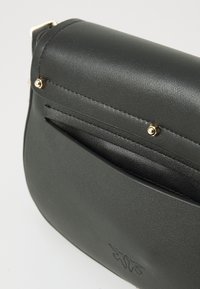 Borsa a tracolla in pelle nera con una tasca frontale sottile, dettagli in borchie dorate e un logo impresso in modo discreto sul pannello frontale inferiore.