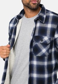 Chemise en flanelle à carreaux bleu marine et blanc avec une doublure intérieure grise, dotée d'une poche à boutons et d'un col pointu. Design décontracté avec une coupe ample.