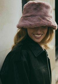 Chapeau seau en fausse fourrure rose avec un large bord, présentant une texture douce et un léger éclat, associé à une veste en cuir noire.