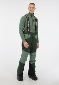 Groene buitenbroeken met verstelbare zwarte banden, versterkte stukken en een contrasterend lichtgroen ontwerp. Beschikt over zakken en duurzaam materiaal.