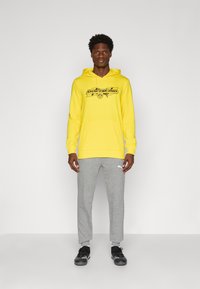 Gul hoodie med logo, frontlomme og snøre; parret med grå sweatsbukser med et lyst logo, iført sorte træningssko.