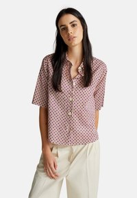 Chemise à manches courtes avec un fond rose, ornée d'un motif géométrique violet et jaune, fermeture boutonnée, col et coupe décontractée.