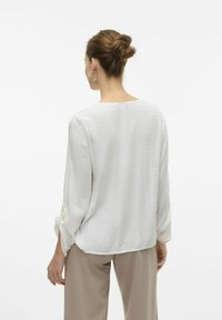 Hvid bluse med prikket tekstur, lange ærmer med opfoldede manchetter og en løs pasform, parret med beige bukser.