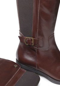 Botte en cuir marron, haute jusqu'au genou, avec des panneaux latéraux élastiques, accent de boucle dorée, texture lisse et semelle noire plate. La vue en quart montre les détails de conception.