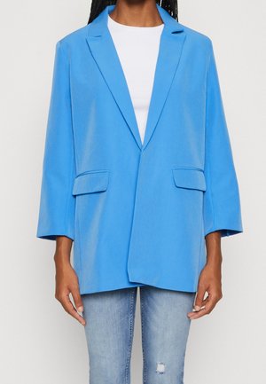 Vrouw draagt een felblauwe oversized blazer met klepzakken over een wit topje en lichtblauwe gescheurde spijkerbroek.