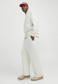 Hanorac alb cu buzunar frontal, purtat cu pantaloni de trening asortați și pantofi bej. Căciulă roșie purtată cu spatele. Material neted și croială lejeră.