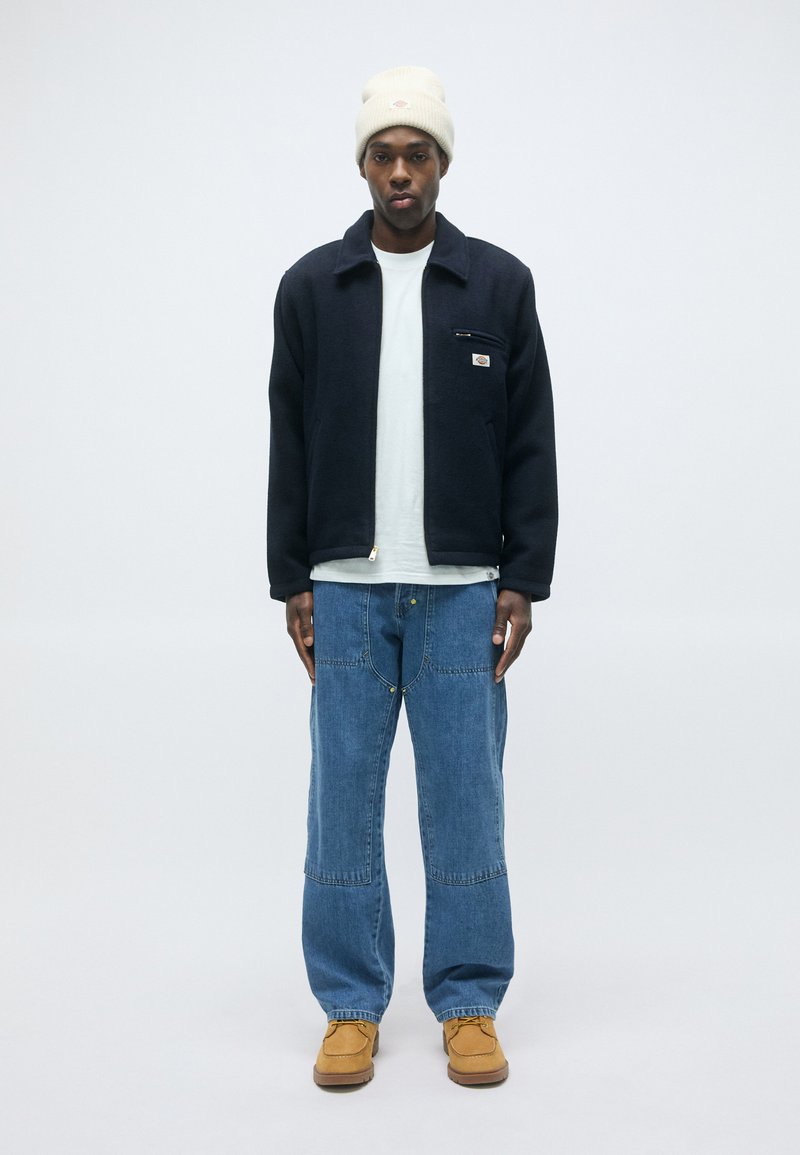 Man staat tegen een effen achtergrond en draagt een witte beanie, een marineblauwe zip-jack, een wit T-shirt, een spijkerbroek in een losse pasvorm en tan geklede veterschoenen.