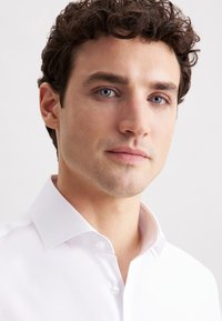 Giovane uomo con capelli castani ricci che indossa una camicia bianca con bottoni, che guarda calmamente la macchina fotografica su uno sfondo chiaro e semplice.