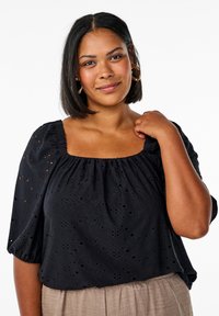 Blusa nera con scollatura quadrata, maniche a sbuffo corte e texture in broderie anglaise. Abbinata a pantaloni chiari marrone con texture.
