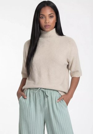 Kobieta o długich czarnych włosach, ubrana w beżowy krótkotkawy sweter z golfem i jasnozielone, paskowane spodnie ze sznurkiem, ręce w kieszeniach.