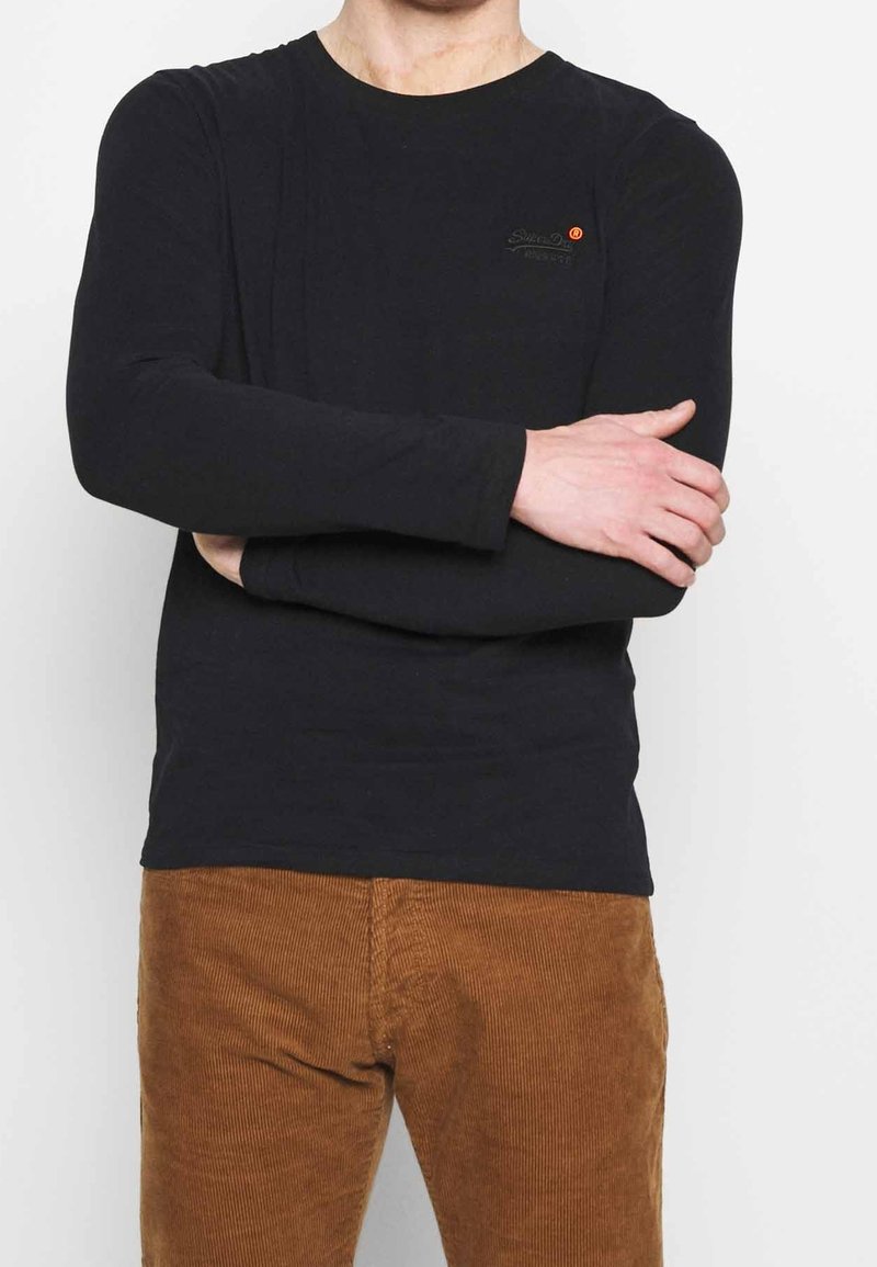 Schwarz langärmliges T-Shirt mit rundem Halsausschnitt und kleinem Logo auf der Brust. Kombiniert mit braunen Cordhosen.