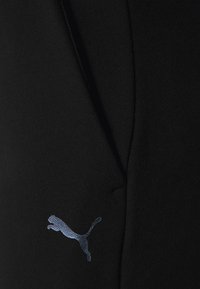 Sudadera negra con una textura suave, que presenta un logotipo de Puma en azul claro en el lado izquierdo. Diseño simple sin patrones ni acentos adicionales.